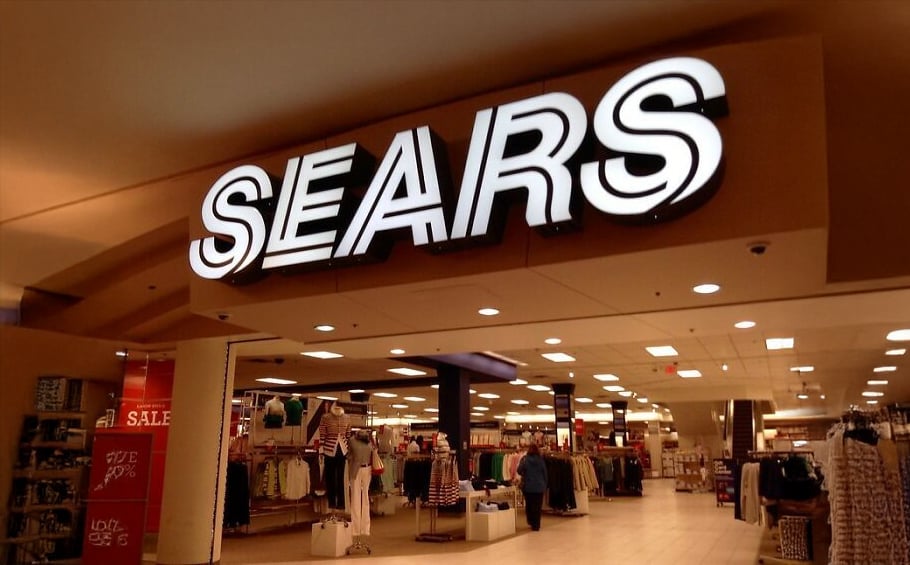 A brightly lit "SEARS" sign hangs above the entrance to a department store, with clothing racks, displays, and a red sale sign visible inside.