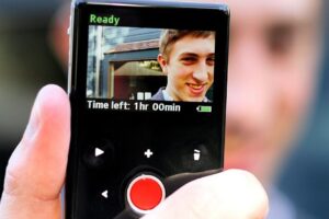 A hand holds a small digital camcorder displaying a young man smiling on its screen. The display reads "Time left: 1hr 00min" and shows the device is ready to record.