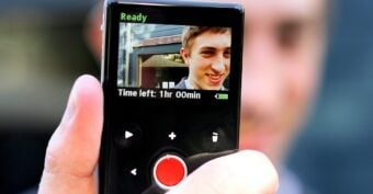 A hand holds a small digital camcorder displaying a young man smiling on its screen. The display reads "Time left: 1hr 00min" and shows the device is ready to record.