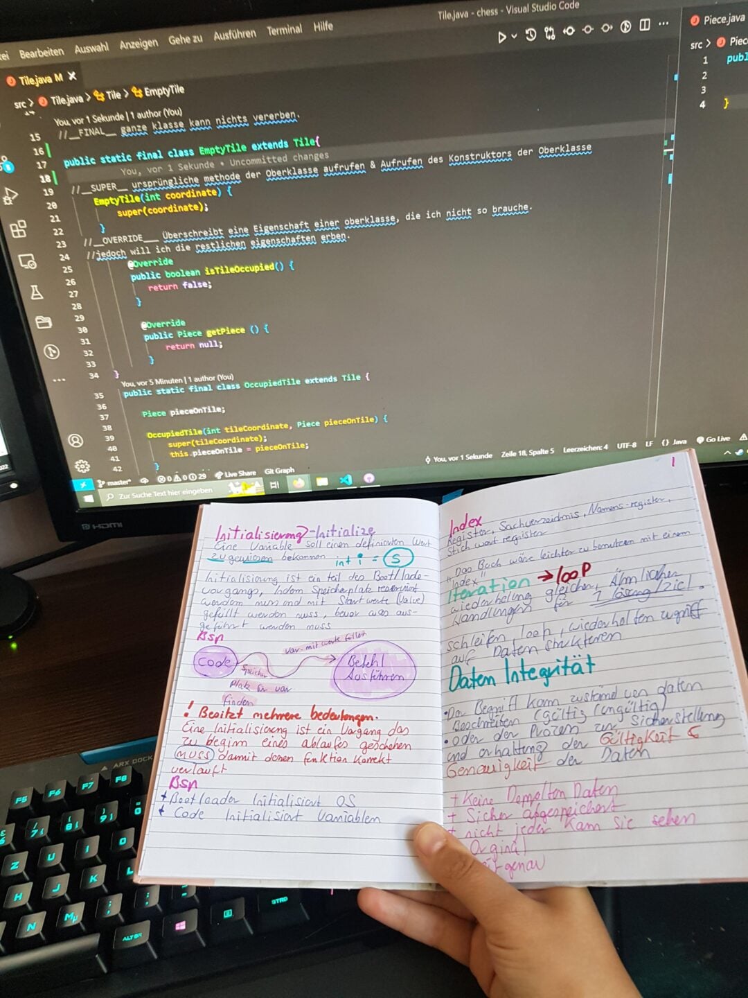 A person holds an open notebook filled with colorful handwritten notes in front of a computer monitor displaying C# code in Visual Studio Code. A keyboard is visible at the bottom.