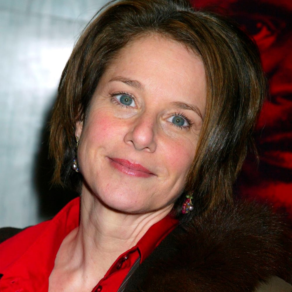 A woman with short brown hair and blue eyes is smiling slightly. She is wearing a red collared shirt and a dark fur-trimmed coat, with dangling earrings, standing in front of a blurred background.