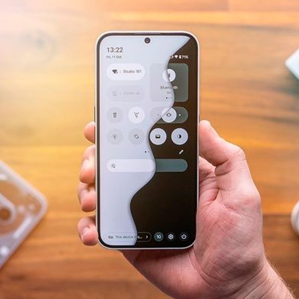 A person holds a smartphone displaying a split screen: the left side is in light mode and the right side is in dark mode. The phone is against a blurred wooden surface background.