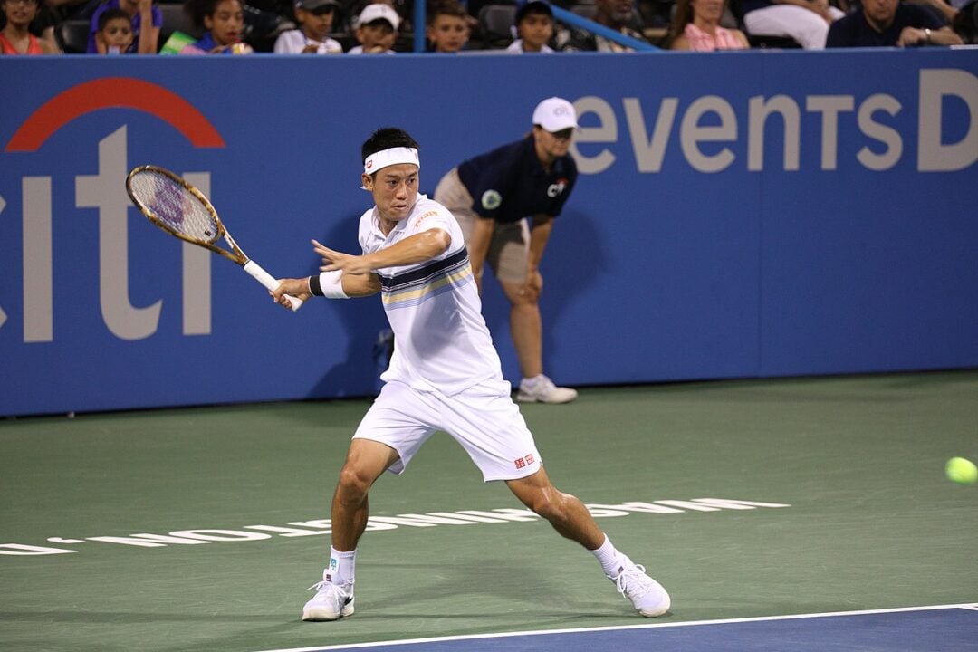 A male tennis player in white sportswear and headband prepares to hit a forehand shot on a hard court during a match, with spectators and a line judge visible in the background.