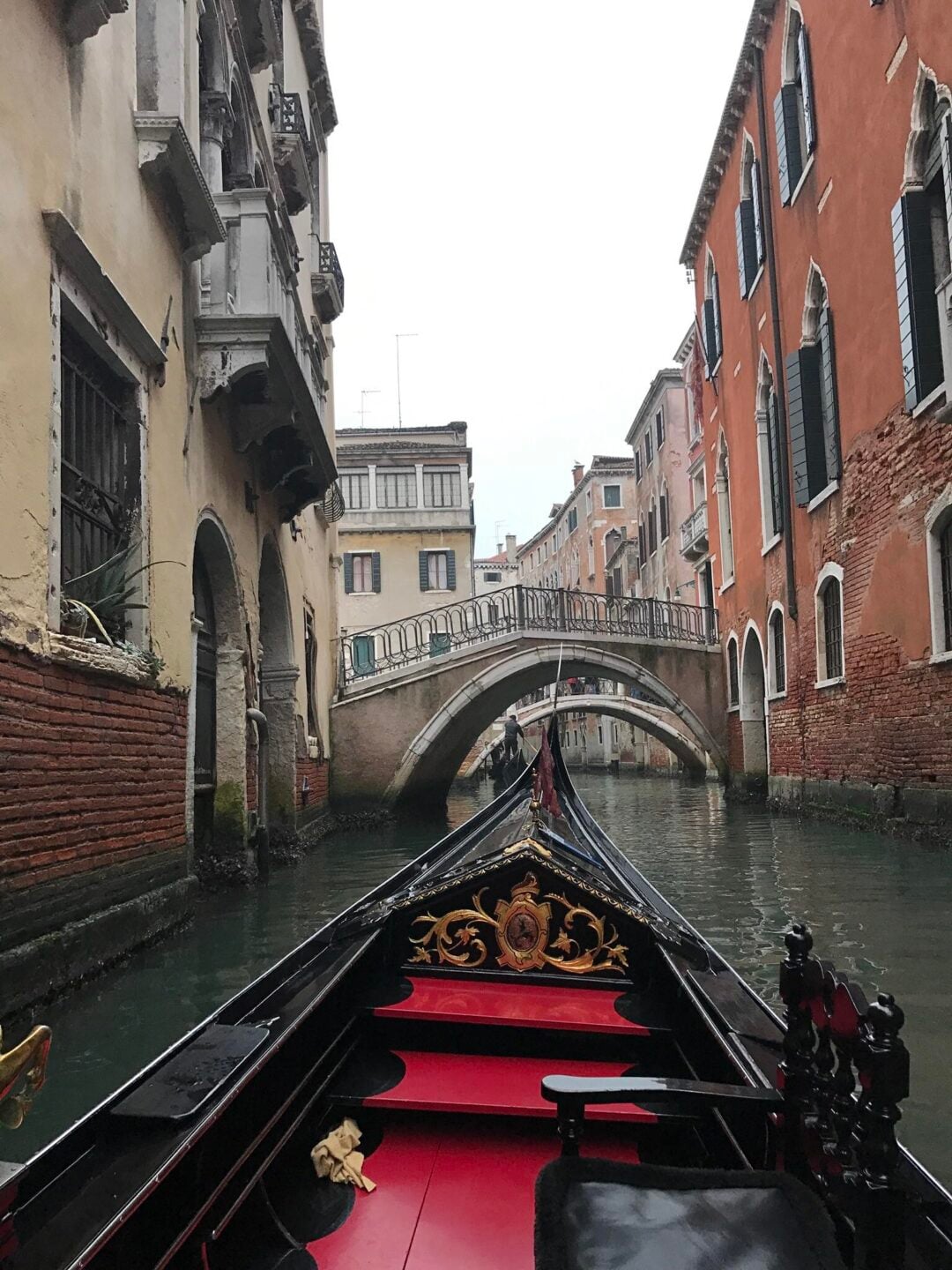 A view from a gondola on a narrow canal in Venice, Italy, lined with old buildings and leading towards a small arched pedestrian bridge.