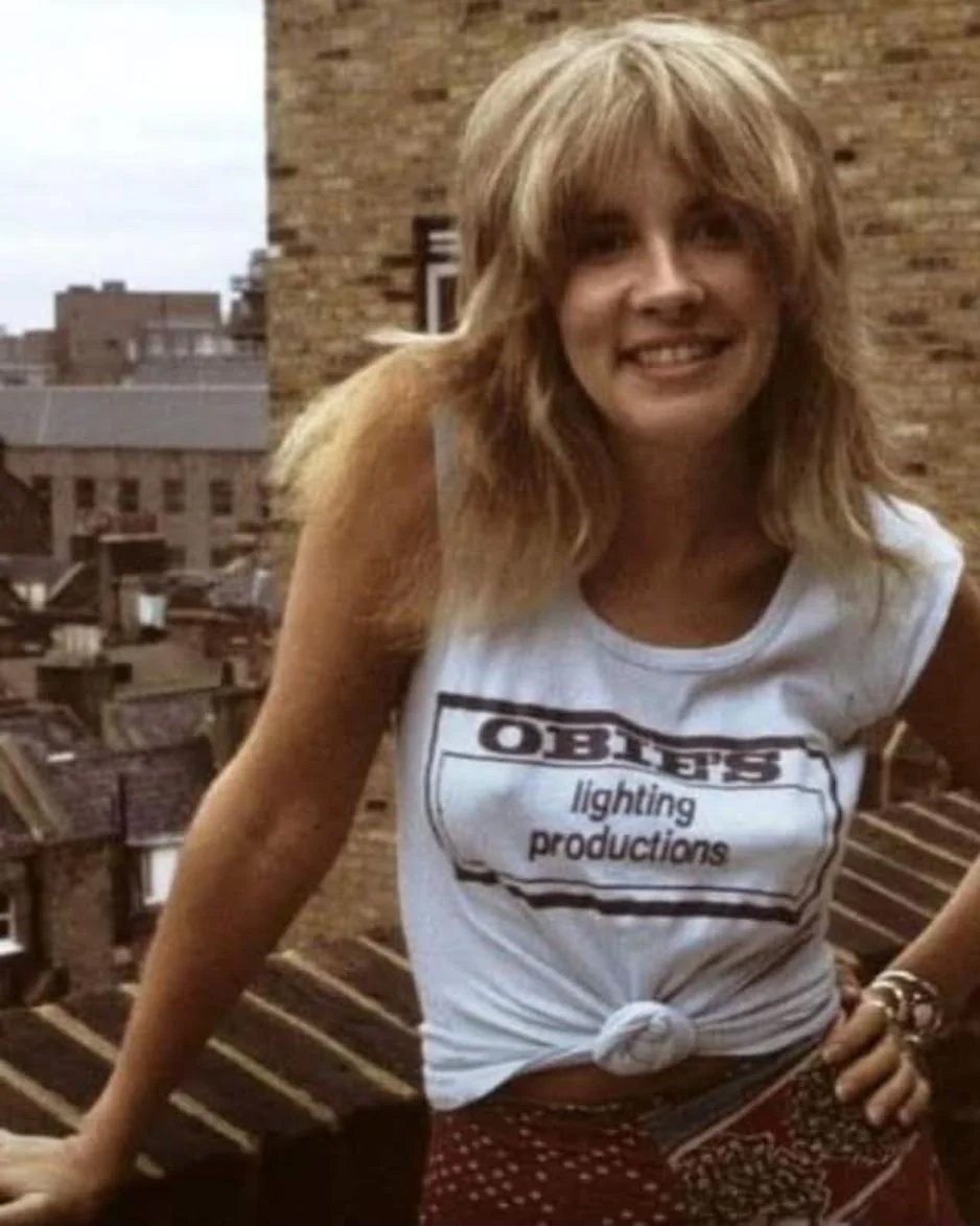 A woman with long, shaggy blonde hair smiles while standing outdoors in front of brick buildings. She wears a light blue sleeveless shirt that reads "OBIES lighting productions" and has it tied in a knot at the waist.