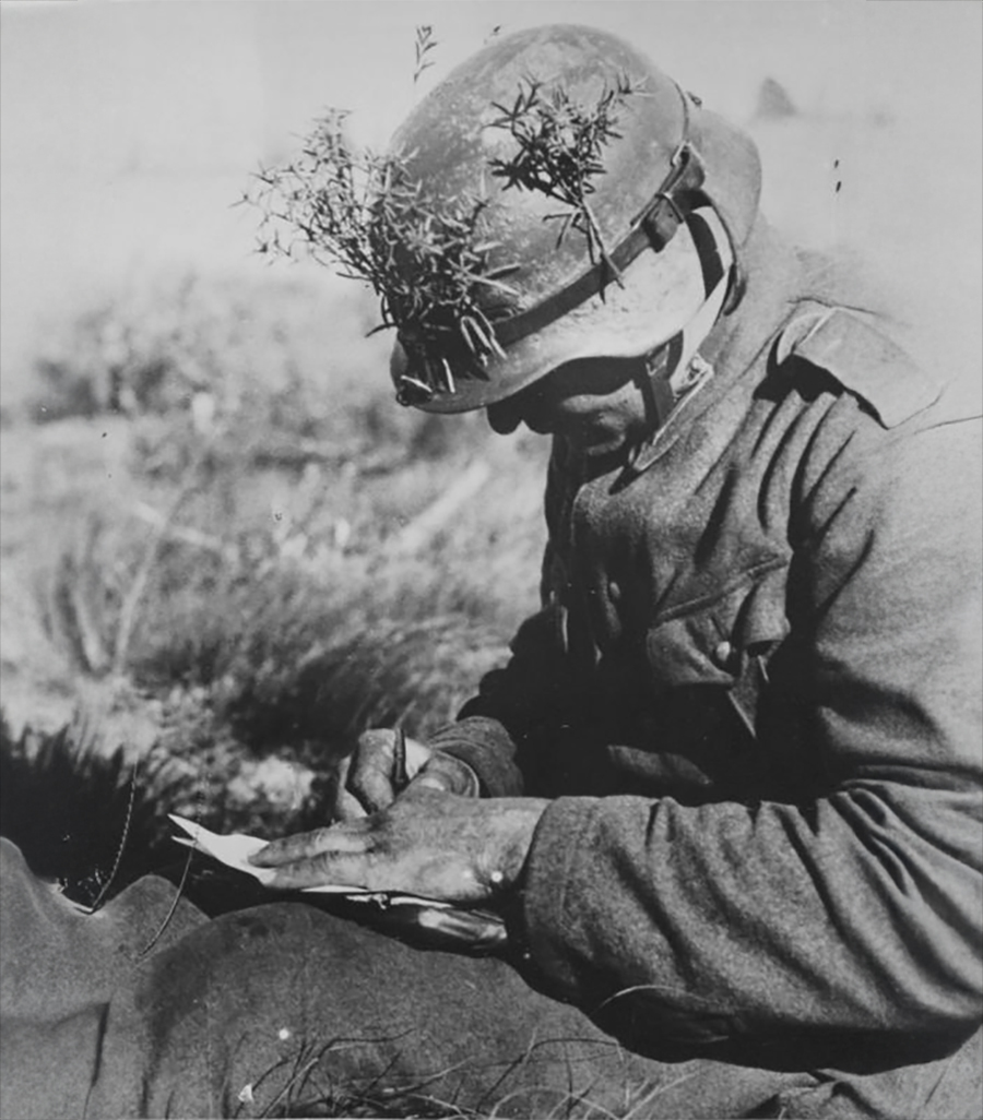 A soldier wearing a helmet with branches for camouflage sits on the ground, looking down while writing on a piece of paper. The scene appears to be outdoors in a grassy area.