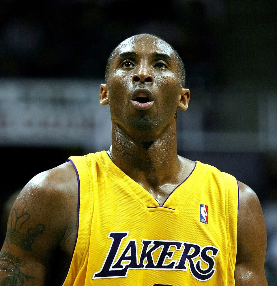 A basketball player in a yellow Lakers jersey stands on the court, looking upward with a focused expression. His skin glistens with sweat under the arena lights.