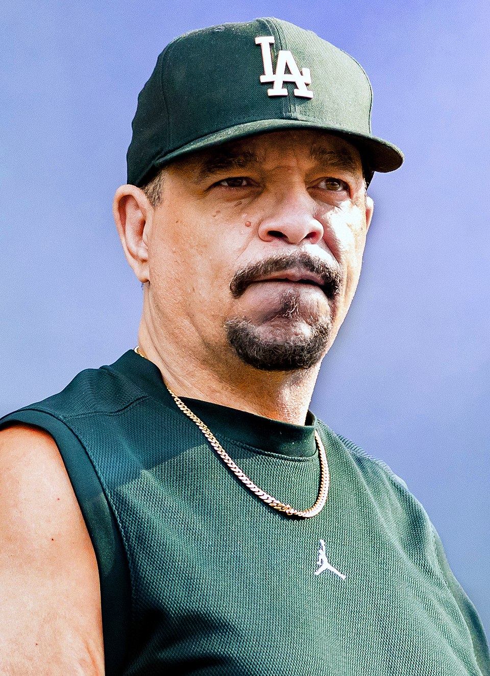 A man wearing a dark green sleeveless shirt, gold chains, and a dark green LA baseball cap stands against a blue, out-of-focus background, looking slightly to the side with a neutral expression.