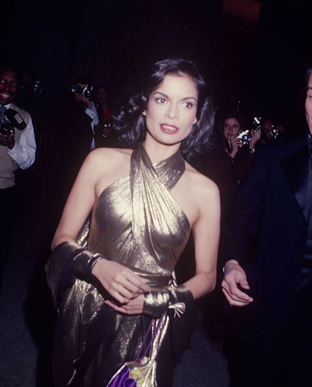 A woman with shoulder-length dark hair wears a shimmering gold one-shoulder dress and matching gloves, holding a small purple bag, as she poses for photos at a formal event. People with cameras surround her in the background.