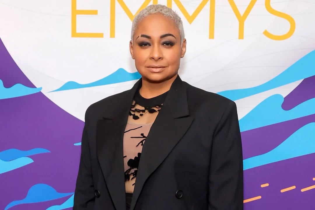 A person with short, light blonde hair wearing a sheer black top and black blazer poses in front of a colorful backdrop with large "EMMYS" text.