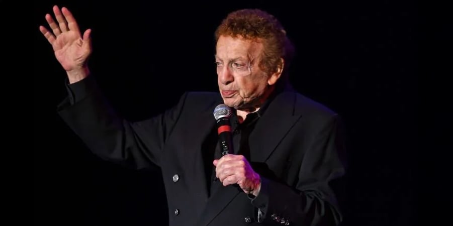 An elderly man in a black suit holds a microphone and raises his left hand while speaking on stage against a dark background.