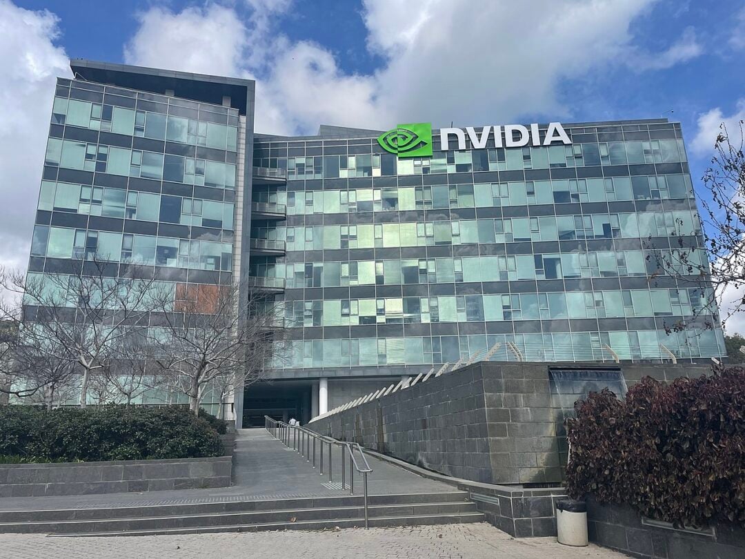 A large modern office building with glass windows displays the NVIDIA logo and green eye symbol near the top. The scene includes a paved walkway, steps, and leafless trees under a partly cloudy sky.