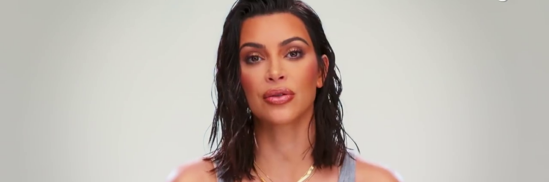 A woman with shoulder-length dark hair, wearing a gray tank top and layered gold necklaces, poses against a plain light background.