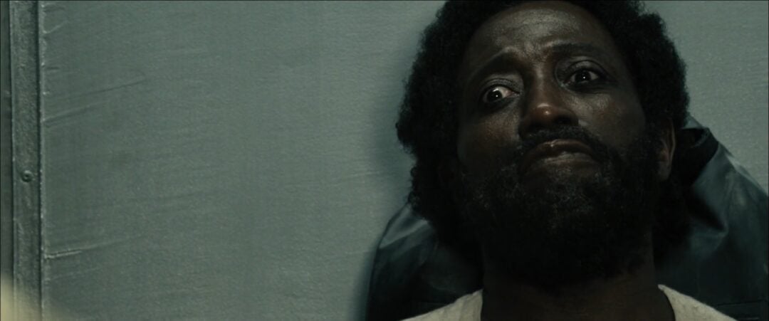 A man with a beard and curly hair leans against a wall, looking upward with wide, worried eyes and a distressed expression. The lighting is dim and the background is plain gray.