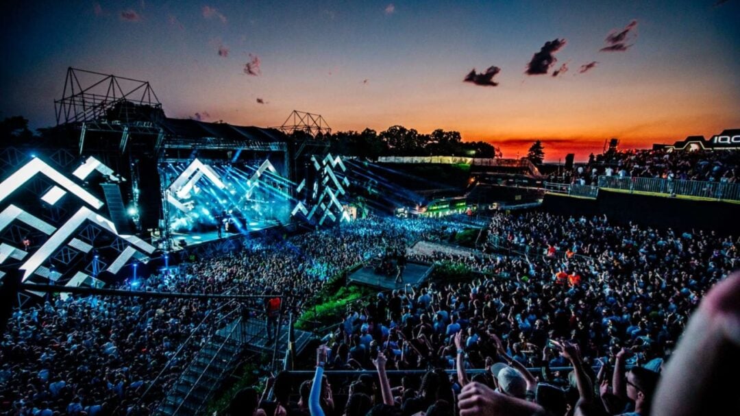 A large crowd gathers at an outdoor music festival at sunset, with colorful stage lights, geometric stage designs, and a vibrant sky in the background. People are cheering and enjoying the lively atmosphere.