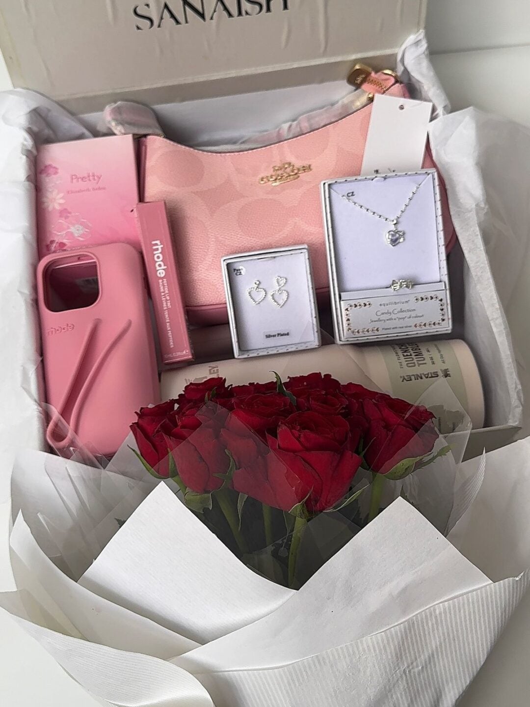 A gift box containing a pink Coach purse, red roses, silver jewelry set with earrings and a necklace, a pink phone case, pink cosmetics, and a gift receipt, surrounded by white tissue paper.