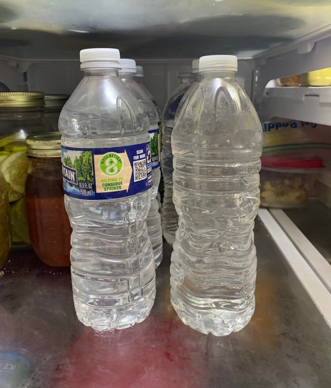 Two plastic water bottles sit side by side inside a refrigerator; one has a label and one does not. Behind them are jars of pickles and other foods. The fridge shelf and other food containers are also visible.