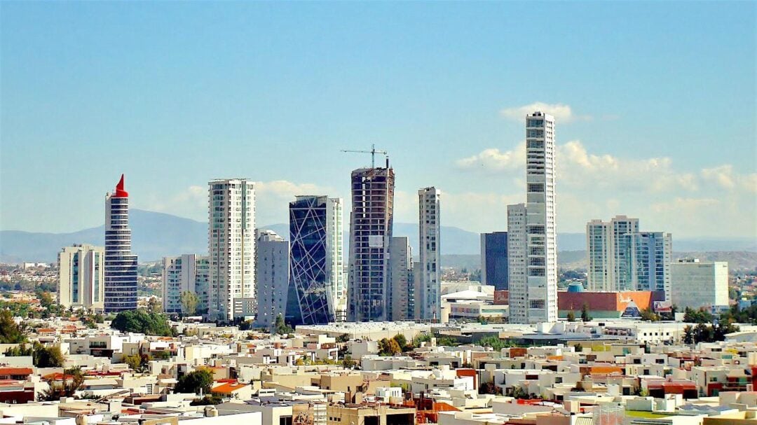 A cityscape featuring a group of modern high-rise buildings with unique architectural designs towering over a densely packed residential area, set against a backdrop of mountains and blue sky.