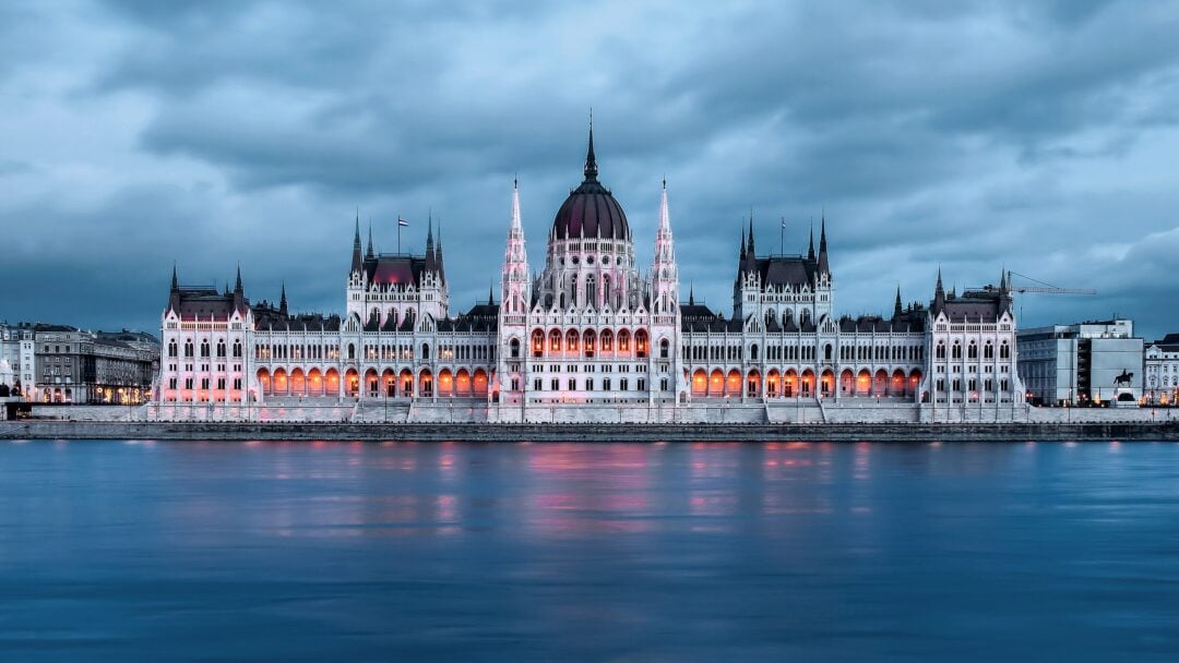 The Hungarian Parliament Building in Budapest is illuminated at dusk, with its reflection visible on the calm Danube River under a cloudy sky.