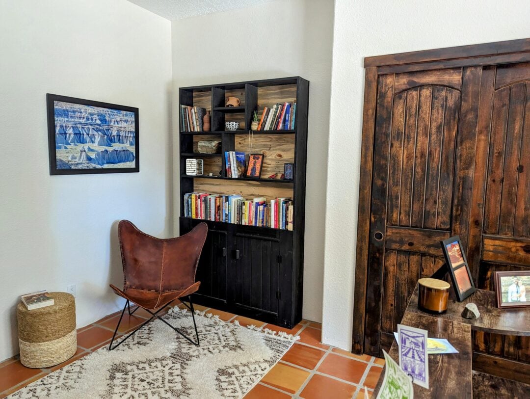 A cozy reading nook with a brown leather butterfly chair, a black bookshelf filled with books, a woven ottoman, a framed landscape photo, a patterned rug, and a wooden cabinet with family photos.