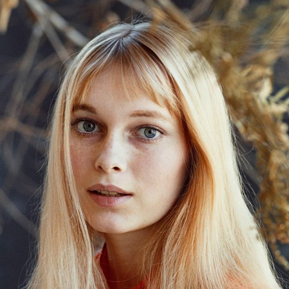 A young woman with long, straight blonde hair and blue eyes gazes softly at the camera. She has a light complexion with freckles and wears a red top. The background is softly blurred with dry branches and neutral tones.