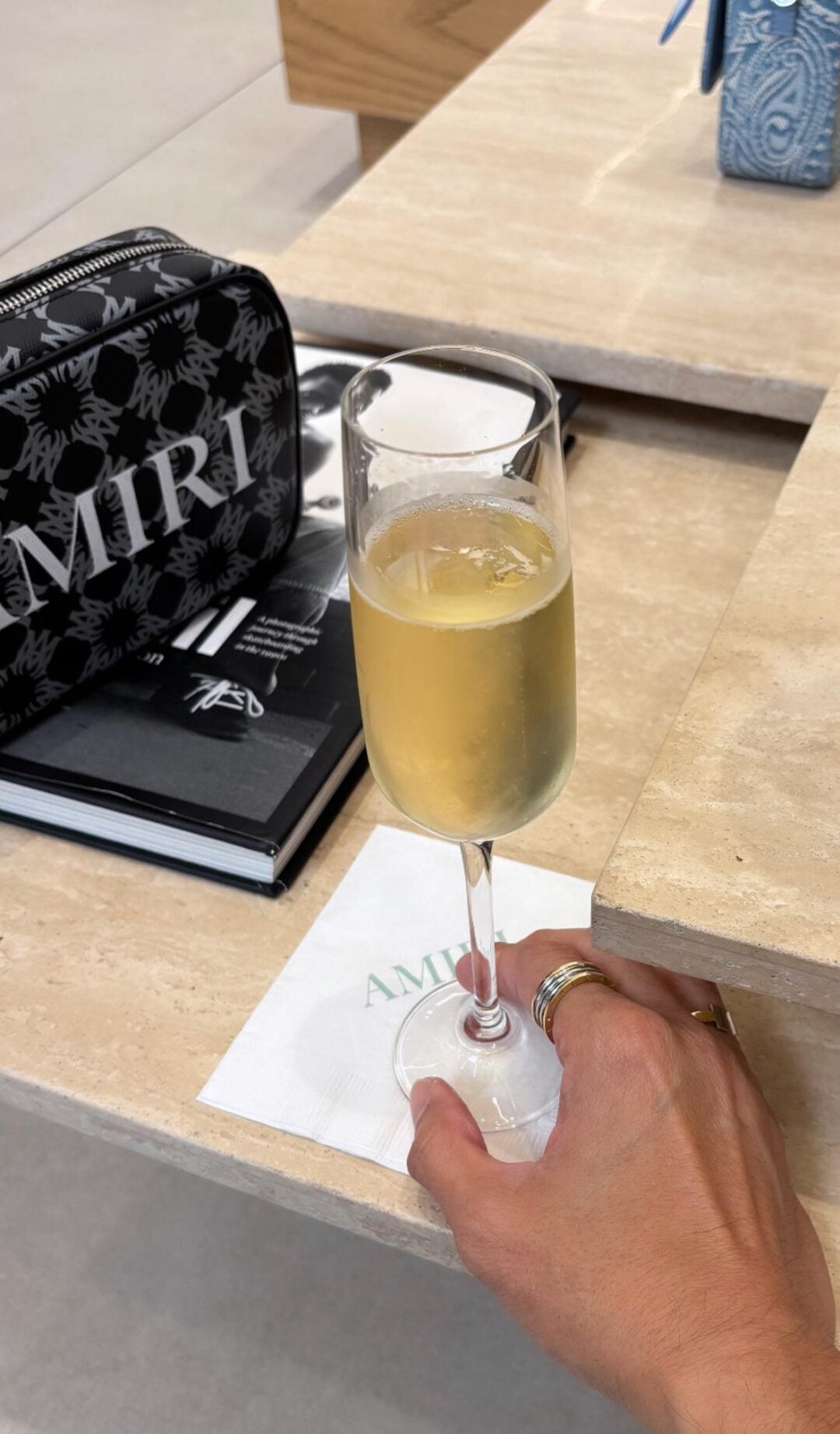 A hand holding a glass of champagne rests on a beige table next to a black AMIRI bag and a stack of magazines. The glass sits on a white napkin with the AMIRI logo.