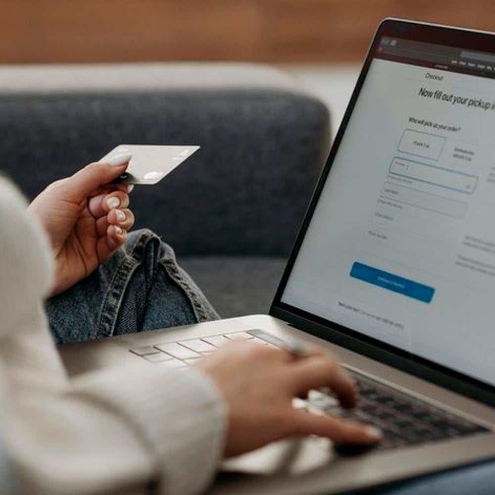 A person holding a credit card in one hand and using the other hand to type on a laptop. The laptop screen displays an online checkout form, suggesting the person is making an online purchase.