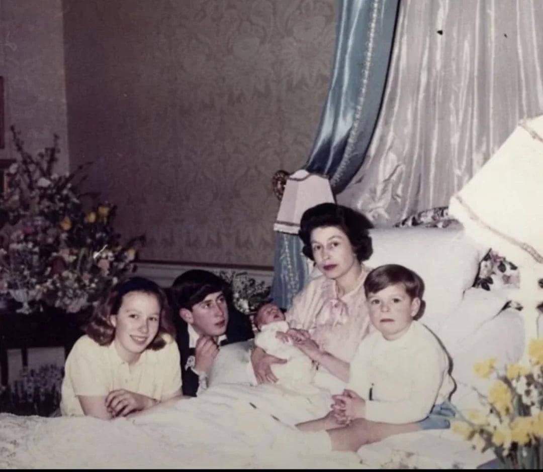 A woman sits up in a canopied bed holding a newborn, surrounded by three children. The room is decorated with flowers and elegant furnishings. The family appears to be posing for a formal photograph.