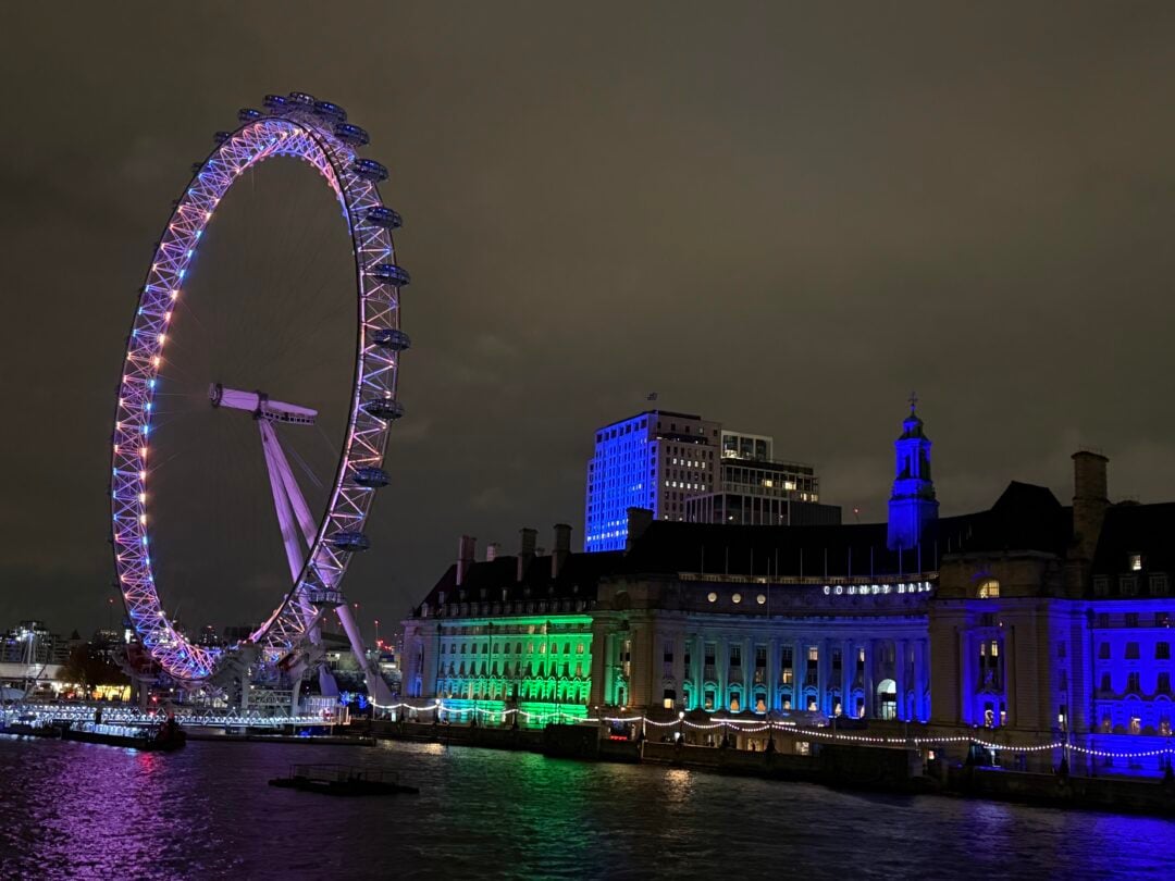 The London Eye illuminated with colorful lights at night, beside the brightly lit riverside County Hall building, reflecting on the River Thames under a cloudy sky.