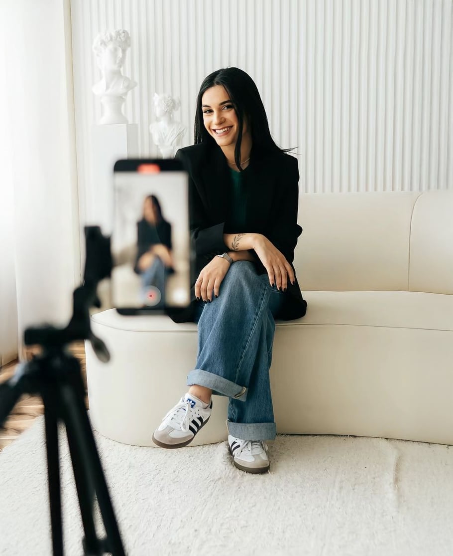 A woman sits on a white sofa, smiling at a smartphone on a tripod filming her. She wears a black blazer, jeans, and white sneakers. The background features white sculptures and a minimalist decor.