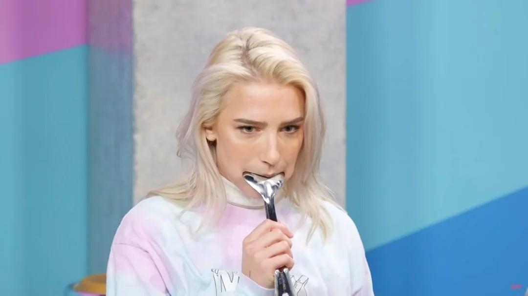 A person with platinum blonde hair wearing a pastel-colored shirt holds a metal wrench in their mouth, standing in front of a colorful blue and purple background.
