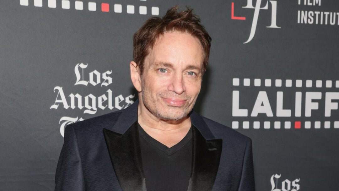 A man with light skin and brown hair, wearing a black suit jacket over a black shirt, smiles at an event with a backdrop featuring the Los Angeles Film Institute and LAIFF logos.