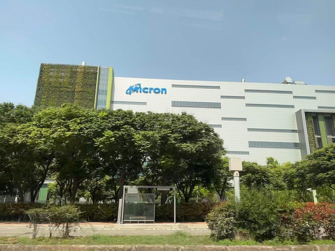A large white building with the "Micron" logo on its facade is partially obscured by rows of green trees and bushes, under a clear sky. A bus stop is visible in front of the building.
