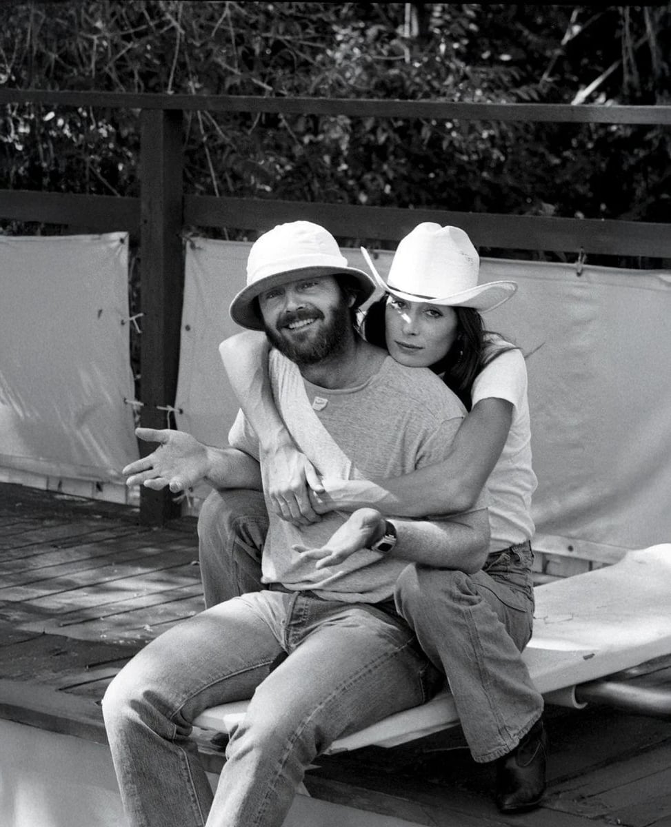 A woman in a cowboy hat hugs a seated man from behind. The man wears a bucket hat and gestures with open hands. They both appear relaxed and are outdoors next to a wooden railing and trees.
