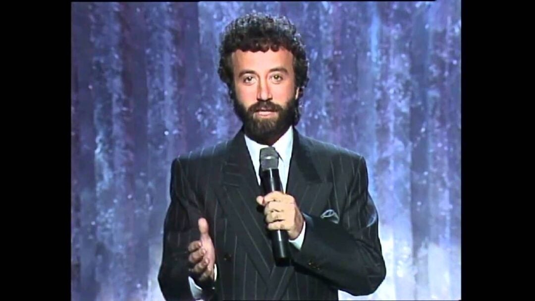 A man with curly dark hair and a beard, wearing a dark pinstripe suit and tie, stands in front of a purple textured background, holding a microphone and gesturing with one hand.