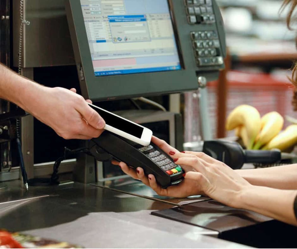 A person is using a smartphone to make a contactless payment at a grocery store checkout, while another person holds the card reader. Bananas are visible on the counter.