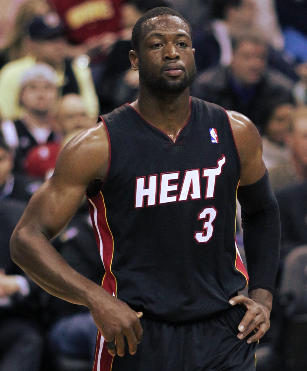 A basketball player in a black Miami Heat jersey with the number 3 stands on a court with hands on hips. Spectators are visible in the blurred background.