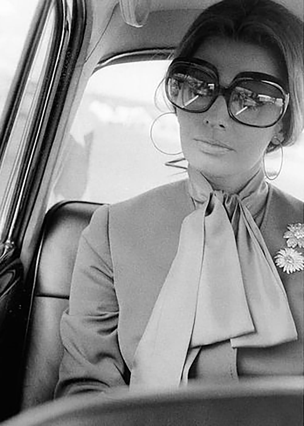 A woman with large sunglasses, hoop earrings, and a bow-tied blouse sits in the back seat of a car, looking out the window. She has a calm expression and wears a floral brooch on her jacket. The image is black and white.