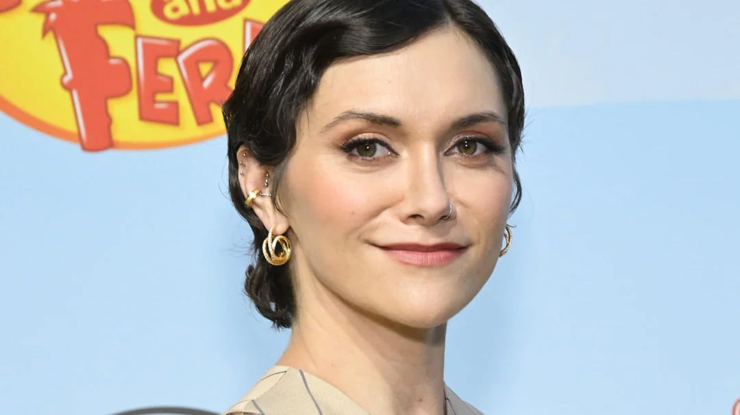 A woman with short, dark wavy hair and gold hoop earrings smiles slightly at the camera. She stands in front of a blue background with part of a colorful cartoon logo visible behind her.