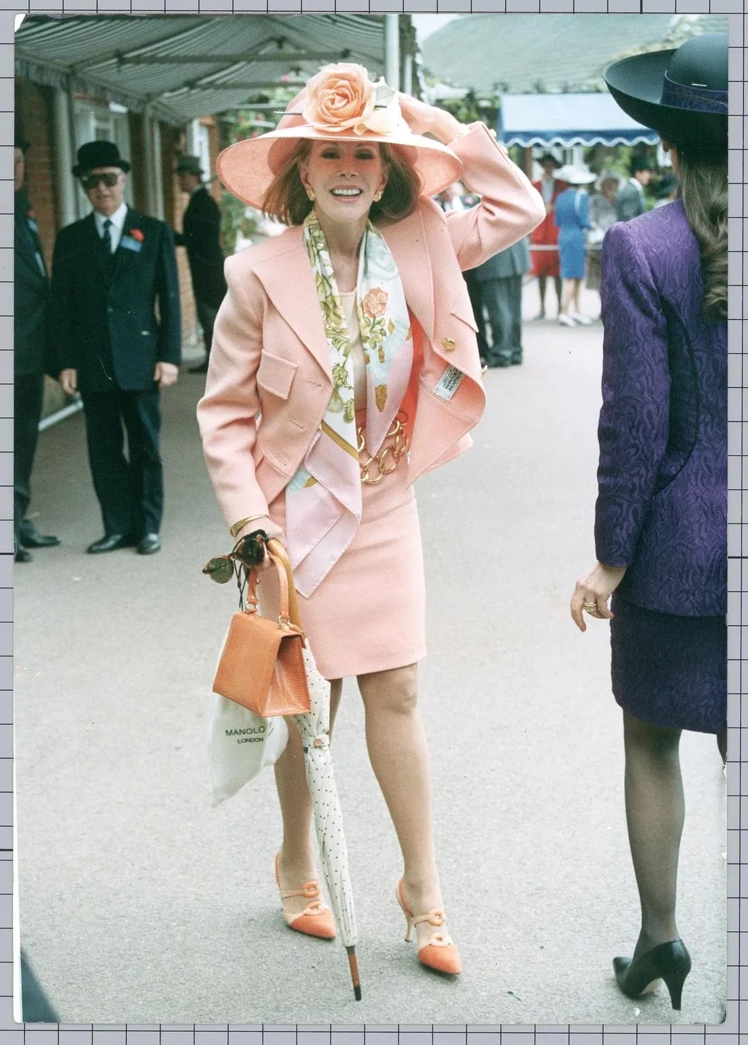 A smiling woman in a pale pink suit, floral scarf, and large pink floral hat stands outdoors holding a handbag and patterned umbrella. She wears matching shoes and is accompanied by another woman in a purple outfit.