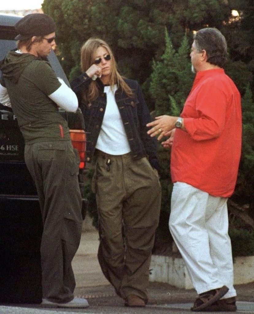 Three people stand outside near a black vehicle and greenery, talking. One woman in sunglasses wipes her eye, while one man wears a red shirt and another man wears a dark hat and sunglasses.