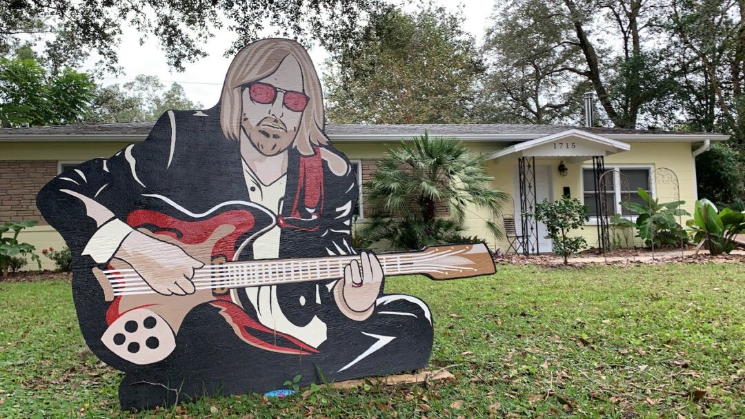 A large painted wood cutout of a man playing an electric guitar is displayed in the front yard of a yellow house with white trim and a few palm trees. The cutout features sunglasses and long hair.