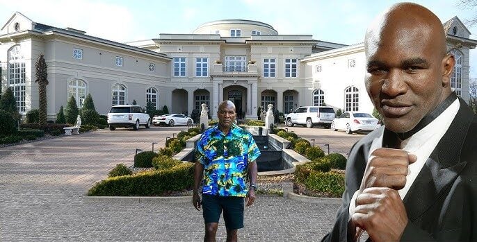 A large mansion with fountains and parked cars in front. A man stands on the driveway wearing a colorful shirt and shorts. An image of the same man in a suit is superimposed. Text reads: “Evander Holyfield’s Former Atlanta Mansion.”