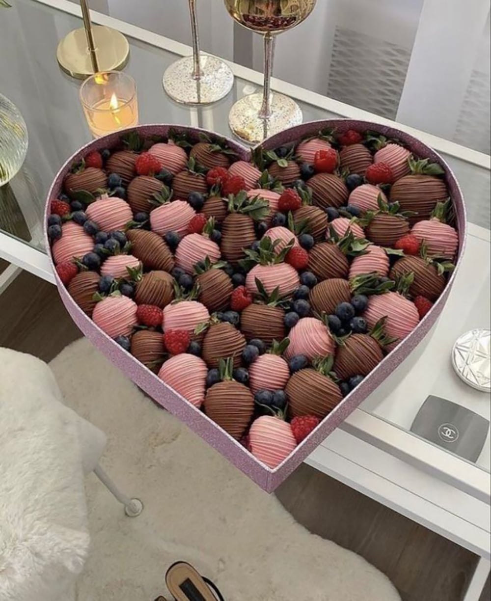 A heart-shaped box filled with chocolate-covered strawberries in pink and brown chocolate, decorated with fresh blueberries and raspberries, placed on a white desk near candles and a decorative tray.