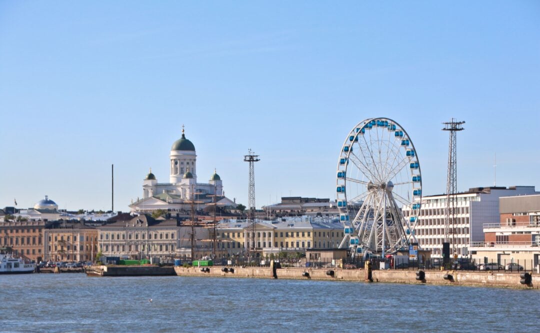 A waterfront cityscape with a large white cathedral, a Ferris wheel, and colorful buildings along the shoreline under a clear blue sky.