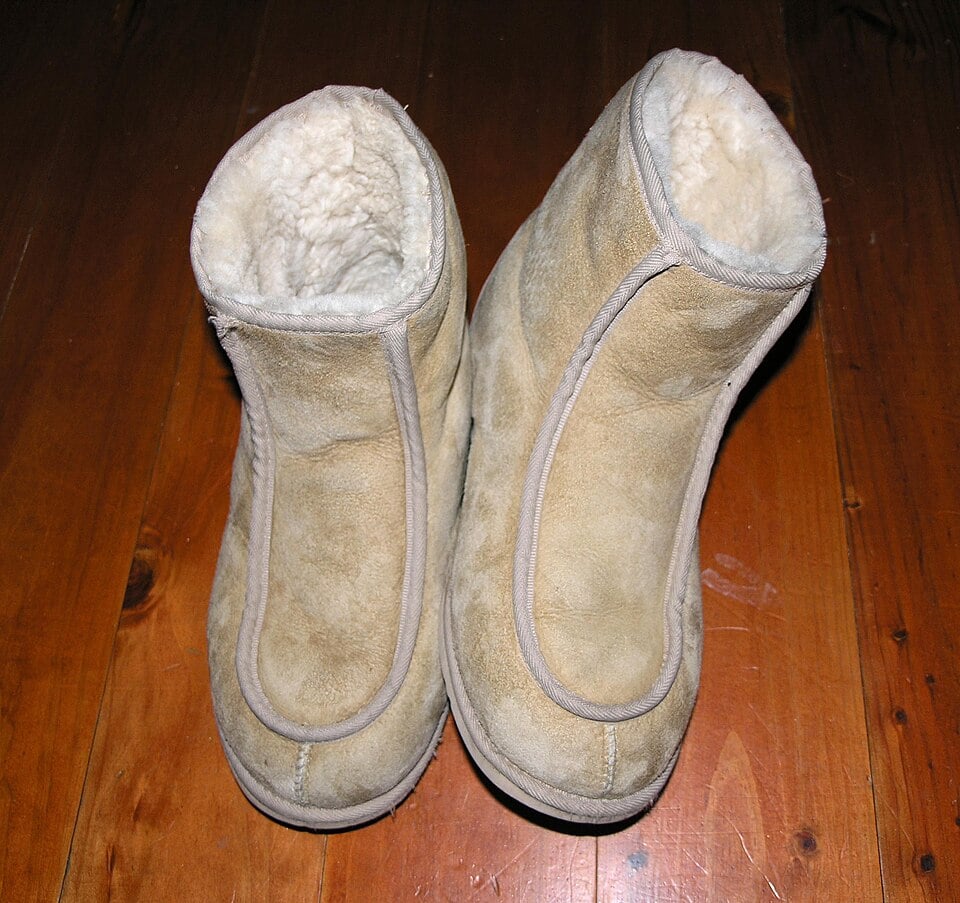 A pair of light beige, suede, fleece-lined winter boots placed side by side on a wooden floor, viewed from above. The boots appear cozy and soft inside.
