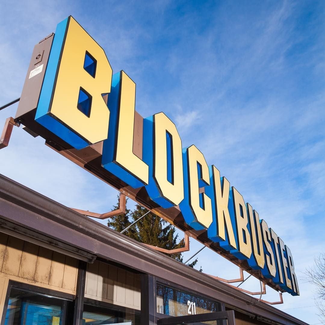 Large yellow and blue BLOCKBUSTER sign on top of a building against a blue sky. The building appears to be an old video rental store.
