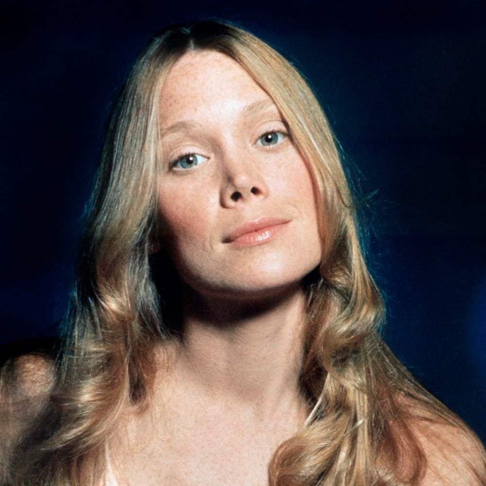 A young woman with long, wavy blonde hair and fair skin looks directly at the camera with a calm expression. She is set against a dark blue background.