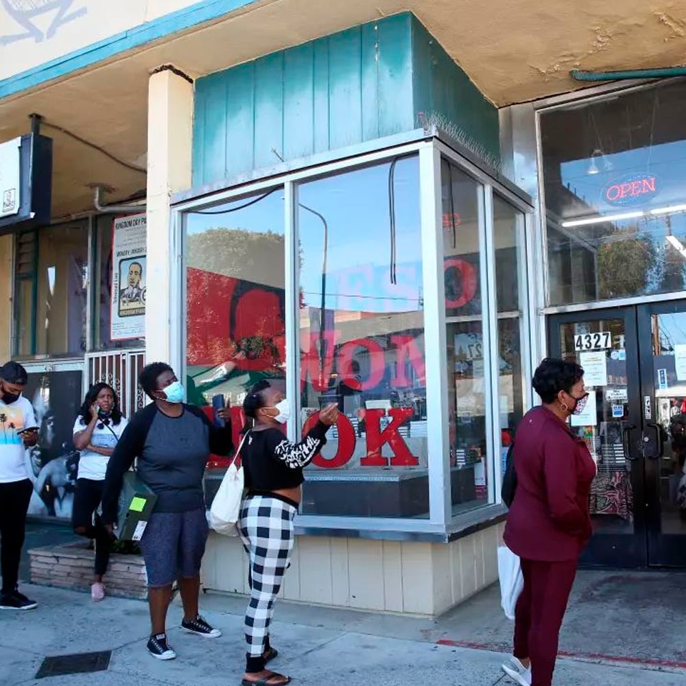People wearing masks stand in line, spaced apart, outside a storefront with large windows and an "OPEN" sign above the door. The sidewalk and part of the building's awning are visible.