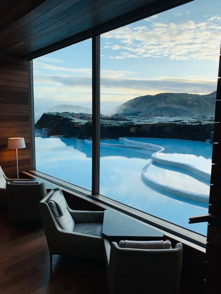 Two modern armchairs and a small table sit by a large window overlooking a serene, steaming blue geothermal pool set against a rocky, mountainous landscape under a partly cloudy sky.
